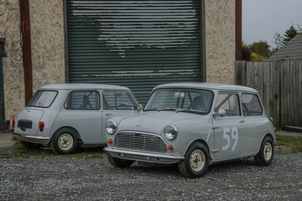 Two pale blue/grey Austin Mini cars parked outside of a garage. One has racing number '59' painted on the side. 